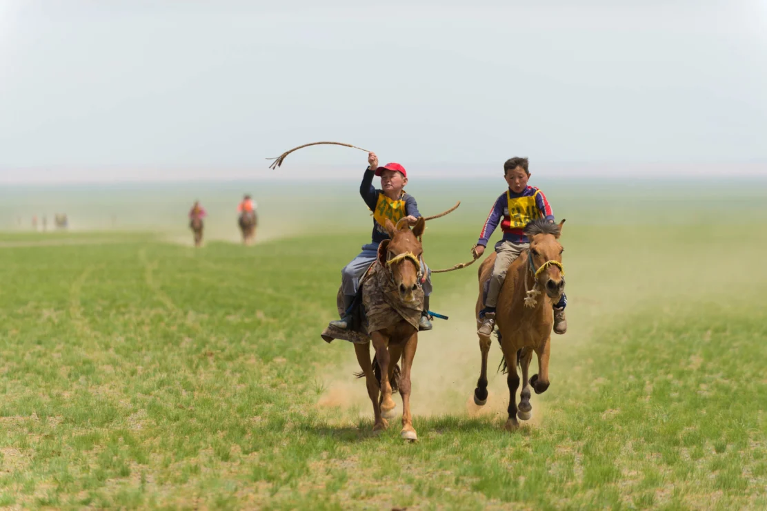 Two people on horseback race across a grassy plain, one raising a whip in the air, with two other riders visible in the distance.