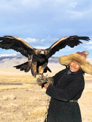 Person holding a large eagle, wearing a fur hat, standing in an open, grassy landscape with distant mountains under a partly cloudy sky.