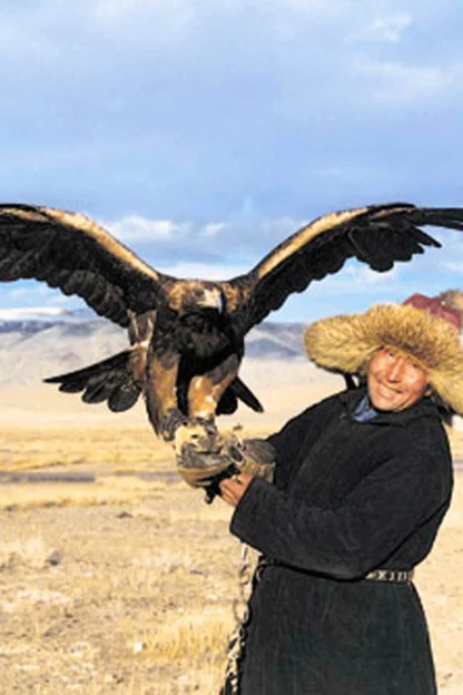 Person holding a large eagle, wearing a fur hat, standing in an open, grassy landscape with distant mountains under a partly cloudy sky.