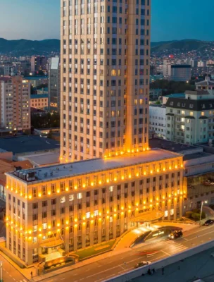 Cityscape with a tall illuminated building surrounded by shorter structures and distant hills under a dusky sky.