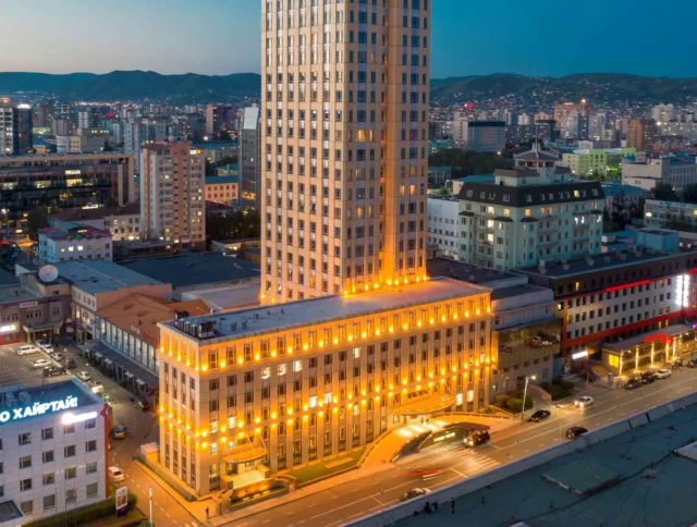 Cityscape with a tall illuminated building surrounded by shorter structures and distant hills under a dusky sky.