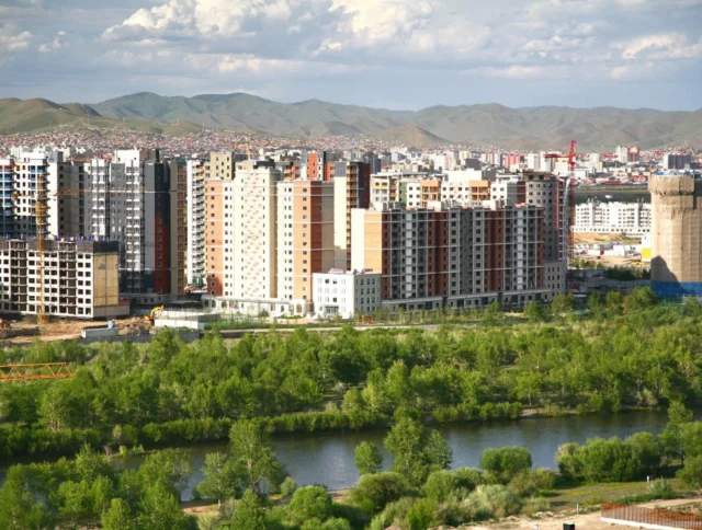 Cityscape with modern apartment buildings, a river, and green trees in the foreground, set against a backdrop of distant hills and a cloudy sky.