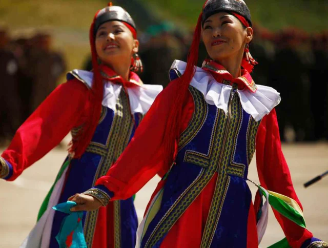 Two performers in traditional, colorful costumes dance outdoors, with blurred figures in the background.