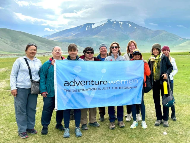 A group of women stand outdoors holding a banner that reads "adventure women." A mountain landscape is visible in the background.