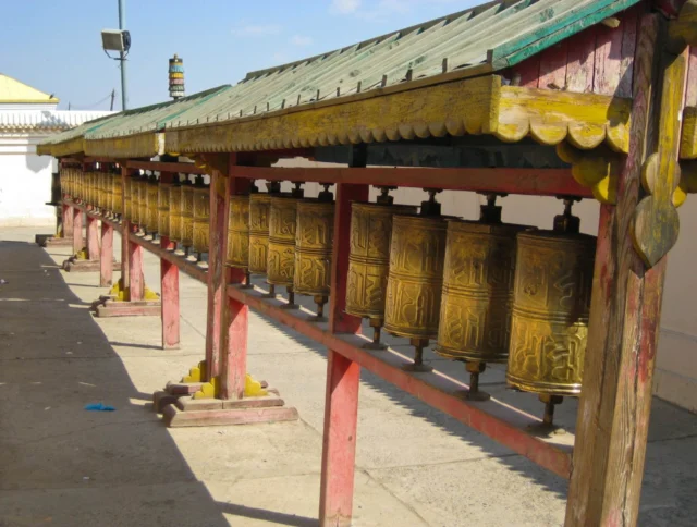 A row of golden prayer wheels under a wooden, green-roofed structure, set against a stone-paved courtyard with a clear blue sky overhead.