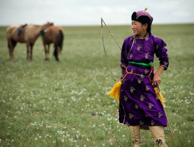 Person in traditional attire holding a bow stands in a grassy field, with two saddled horses in the background.