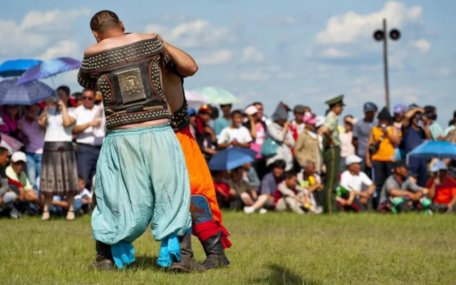 Two men in traditional attire wrestling outdoors, with a crowd watching in the background under blue skies.