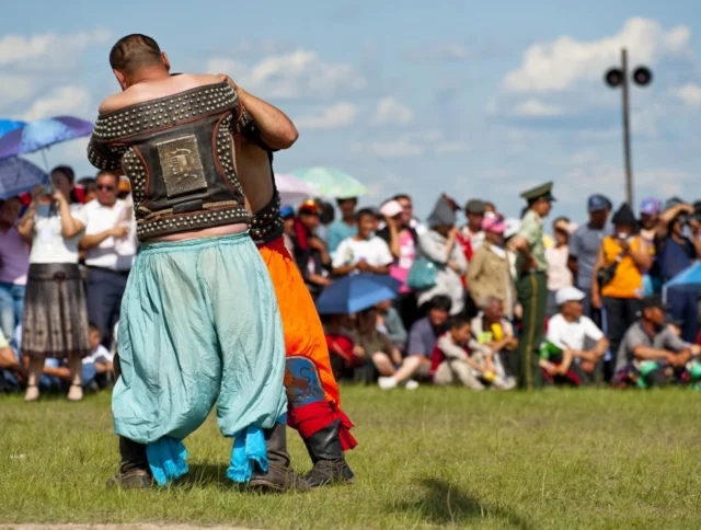 Two men in traditional attire wrestling outdoors, with a crowd watching in the background under blue skies.