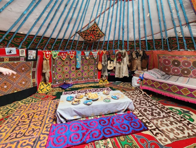 Interior of a decorated yurt with colorful rugs, patterns on walls, musical instruments, and a low table set for a meal.