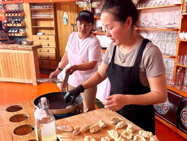 Two women are cooking dumplings in a kitchen. One is wearing a white shirt and cap, stirring a pan, while the other in a black apron arranges dumplings on a counter.