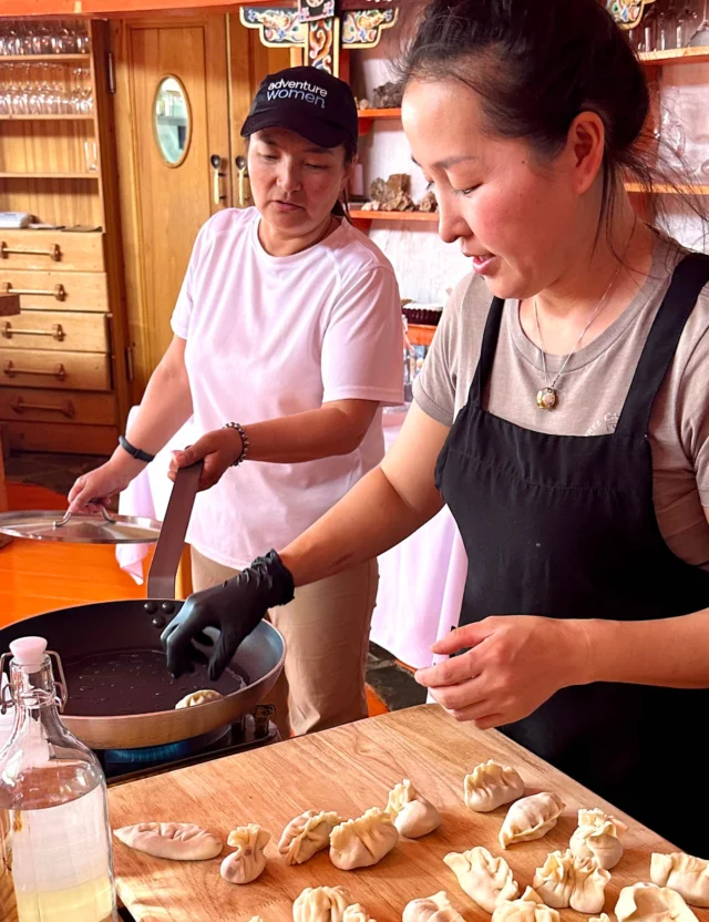 Two women are cooking dumplings in a kitchen. One is wearing a white shirt and cap, stirring a pan, while the other in a black apron arranges dumplings on a counter.