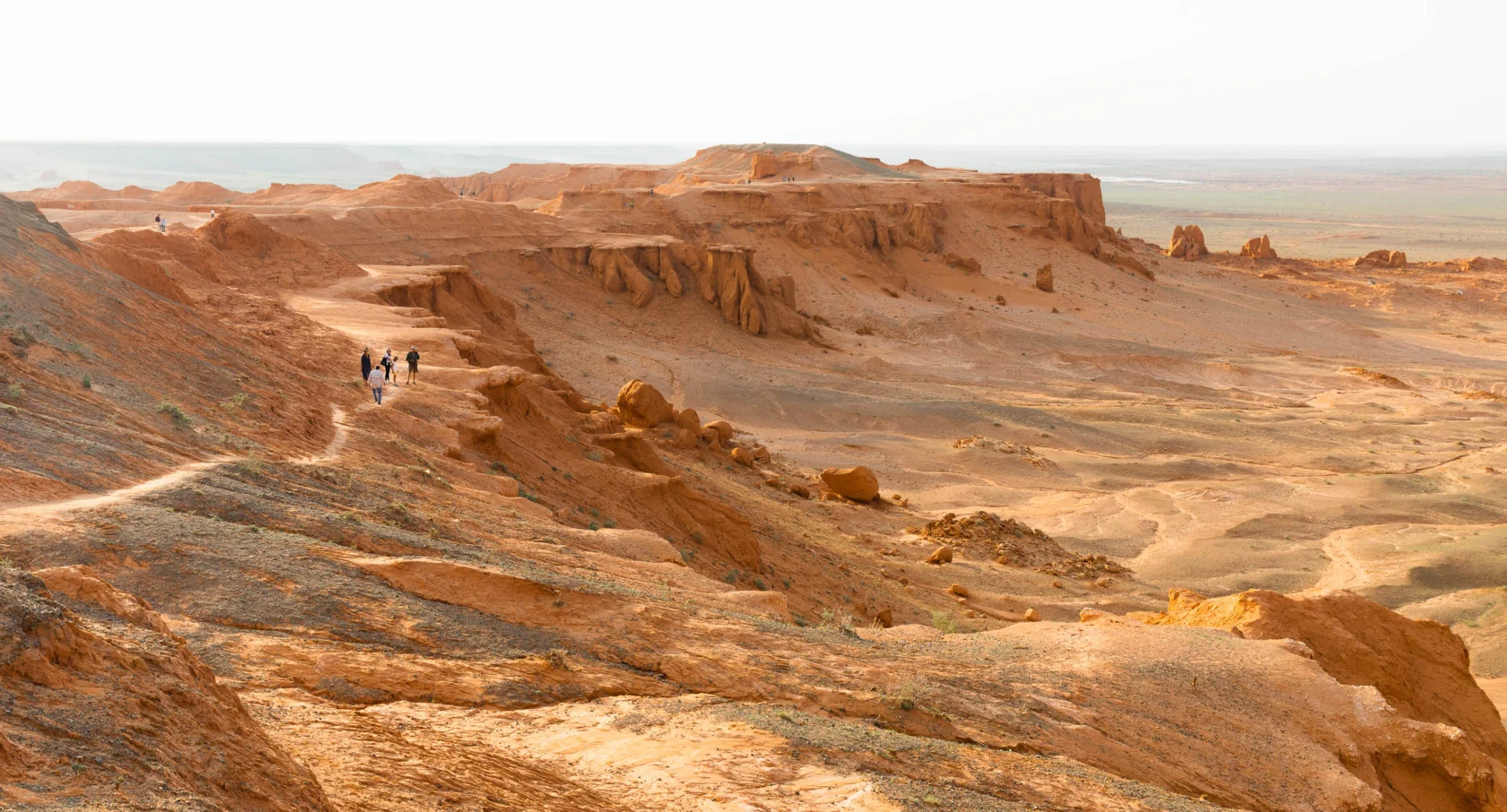 Three people walk along a rocky trail in a vast, arid landscape with sandstone formations and a distant horizon.