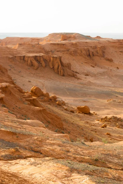 Three people walk along a rocky trail in a vast, arid landscape with sandstone formations and a distant horizon.