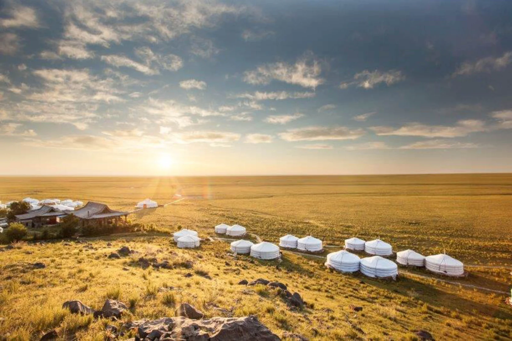 Yurt settlement on a vast grassy plain under a sunrise sky, with rocky foreground and scattered clouds.