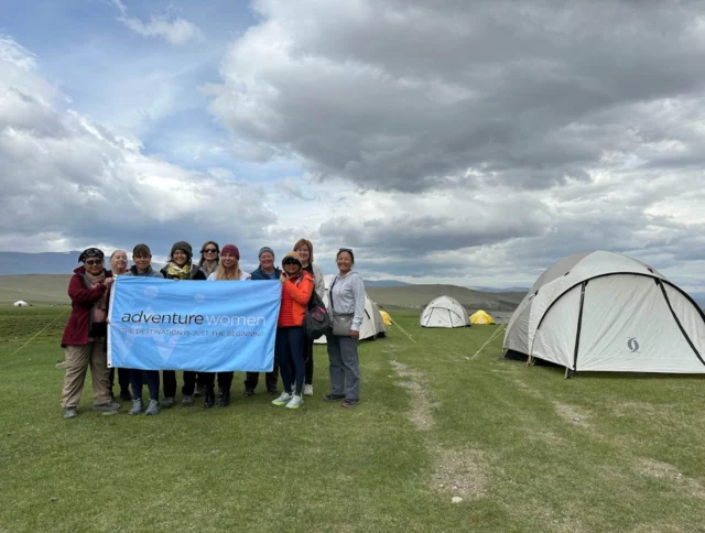 A group of women stand on grassy terrain holding a blue "adventure women" banner. Tents are pitched in the background under a cloudy sky.