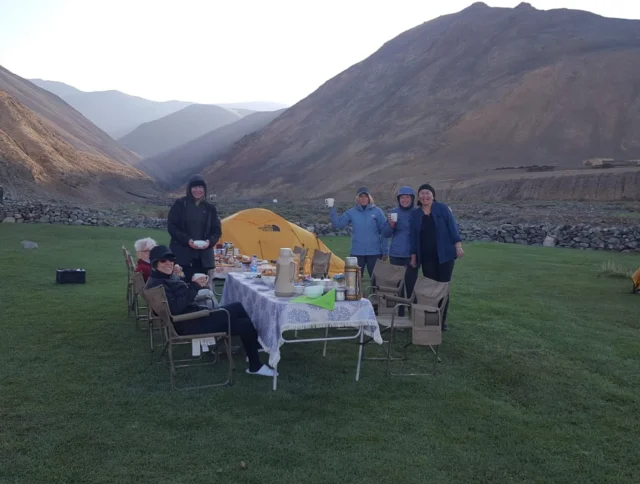 A group of people sits and stands around a table with food, set on green grass among tents and mountains.