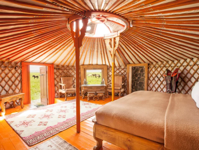 The interior of a yurt features a bed, chairs, a small table, and a rug. The wooden roof structure is visible, and the doorway opens to a grassy landscape.