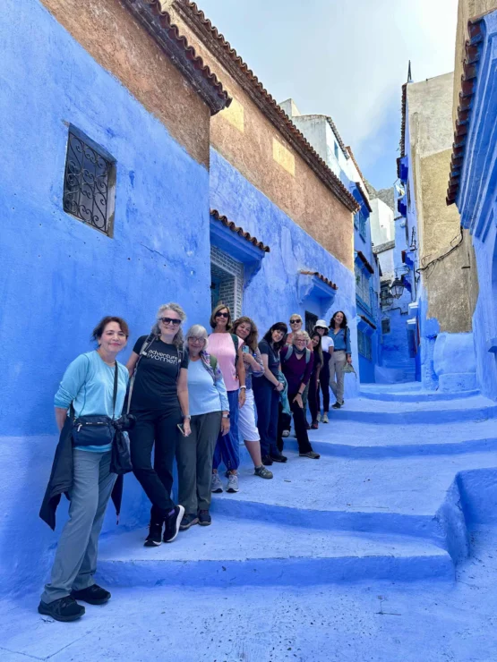 A group of people stands in a line on a street with bright blue walls, in an alleyway with steps.