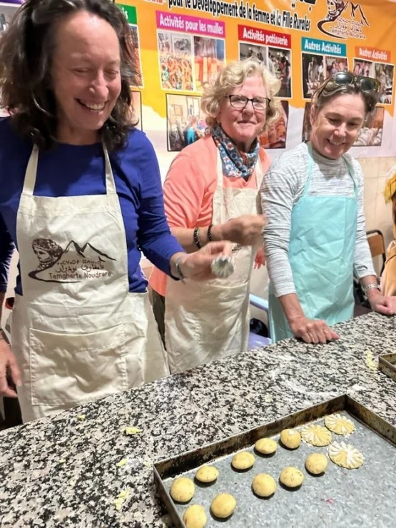 Three women in aprons stand around a kitchen counter, shaping dough on a tray. A colorful poster is visible in the background.