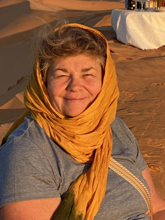 Woman wearing an orange headscarf and gray shirt, smiling in a desert setting with sand dunes and a table in the background.