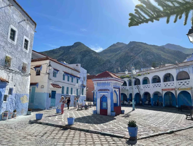 A sunlit square with blue and white buildings, a central pavilion, and mountains in the background. Visitors are walking and exploring the area.