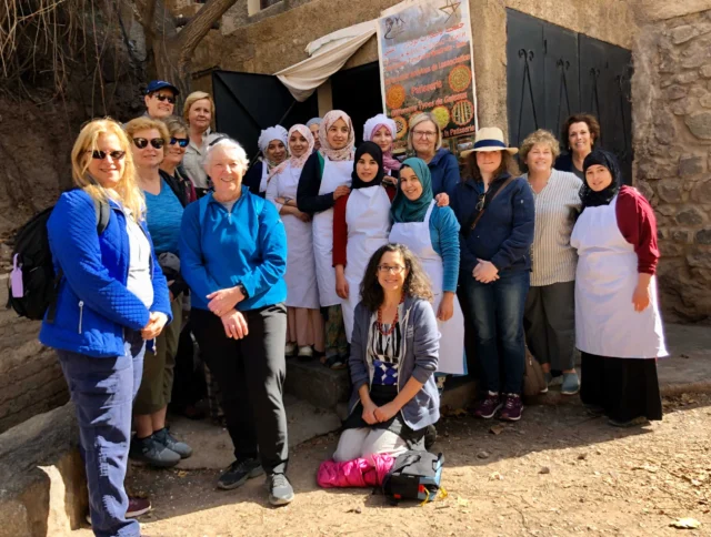 A group of people, including several wearing aprons and headscarves, pose together outside a rustic building with a stone wall.