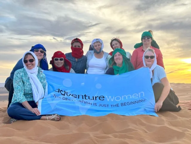 A group of women in headscarves holding a blue "Adventure Women" banner sit on sand dunes at sunset.