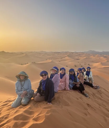 A group of people sit on sand dunes in a desert landscape at sunset, wearing hats and headscarves.