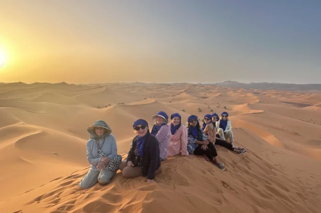 A group of people sit on sand dunes in a desert landscape at sunset, wearing hats and headscarves.