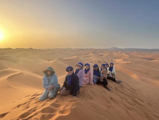 A group of people sit on sand dunes in a desert landscape at sunset, wearing hats and headscarves.