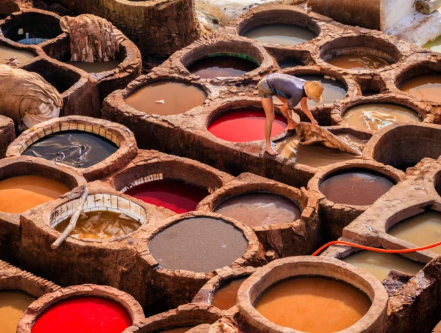 A person works at a traditional tannery with numerous circular pits filled with colorful liquids, likely for dyeing leather.