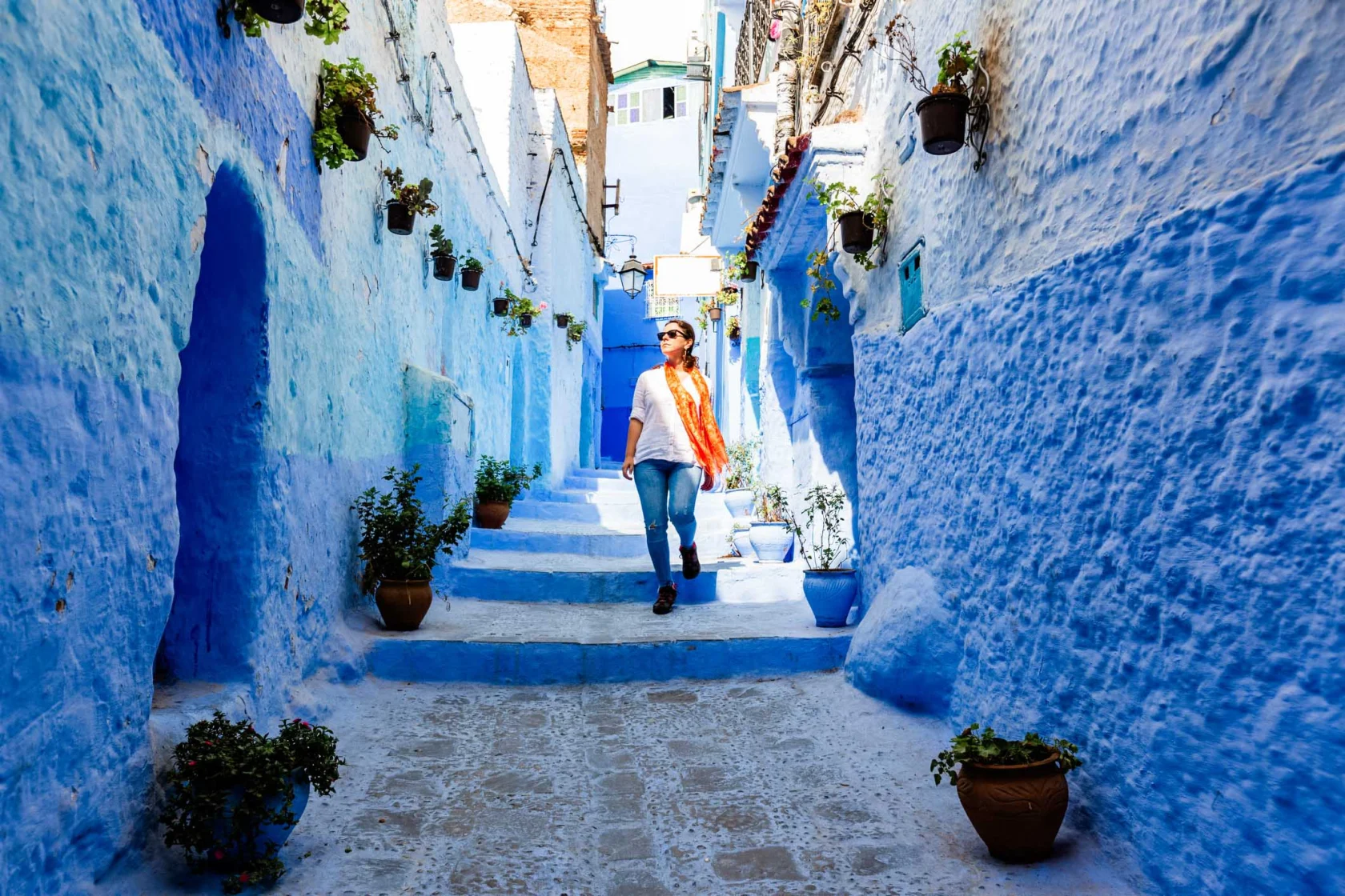 Person walking down a narrow blue-painted street with potted plants lining the walls.