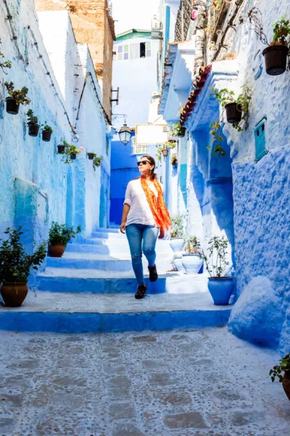 Person walking down a narrow blue-painted street with potted plants lining the walls.