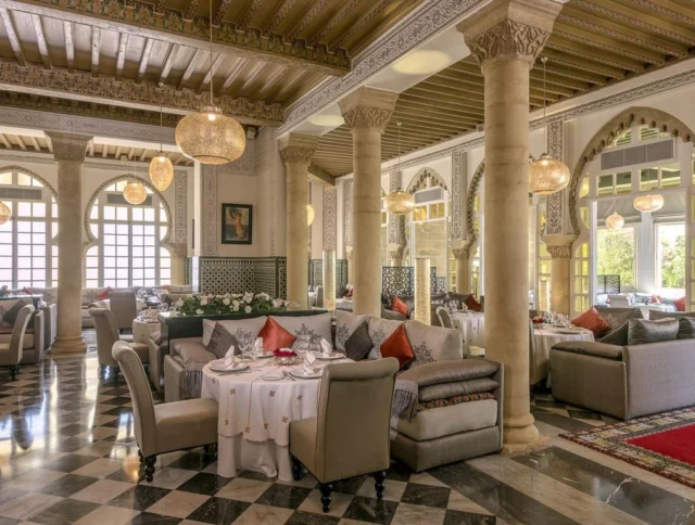 Luxurious restaurant interior with ornate columns, decorative arches, and plush seating. Tables are set with white tablecloths and elegant place settings, under large spherical chandeliers.