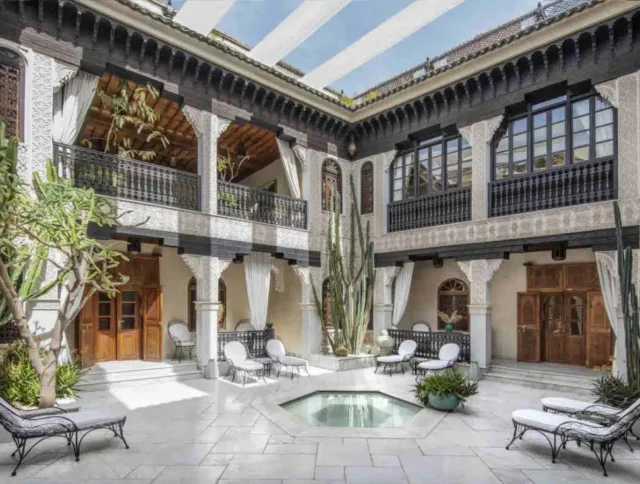 Elegant courtyard with white furniture, central fountain, and surrounding greenery. Wood and wrought iron balconies frame the space, under a striped awning.