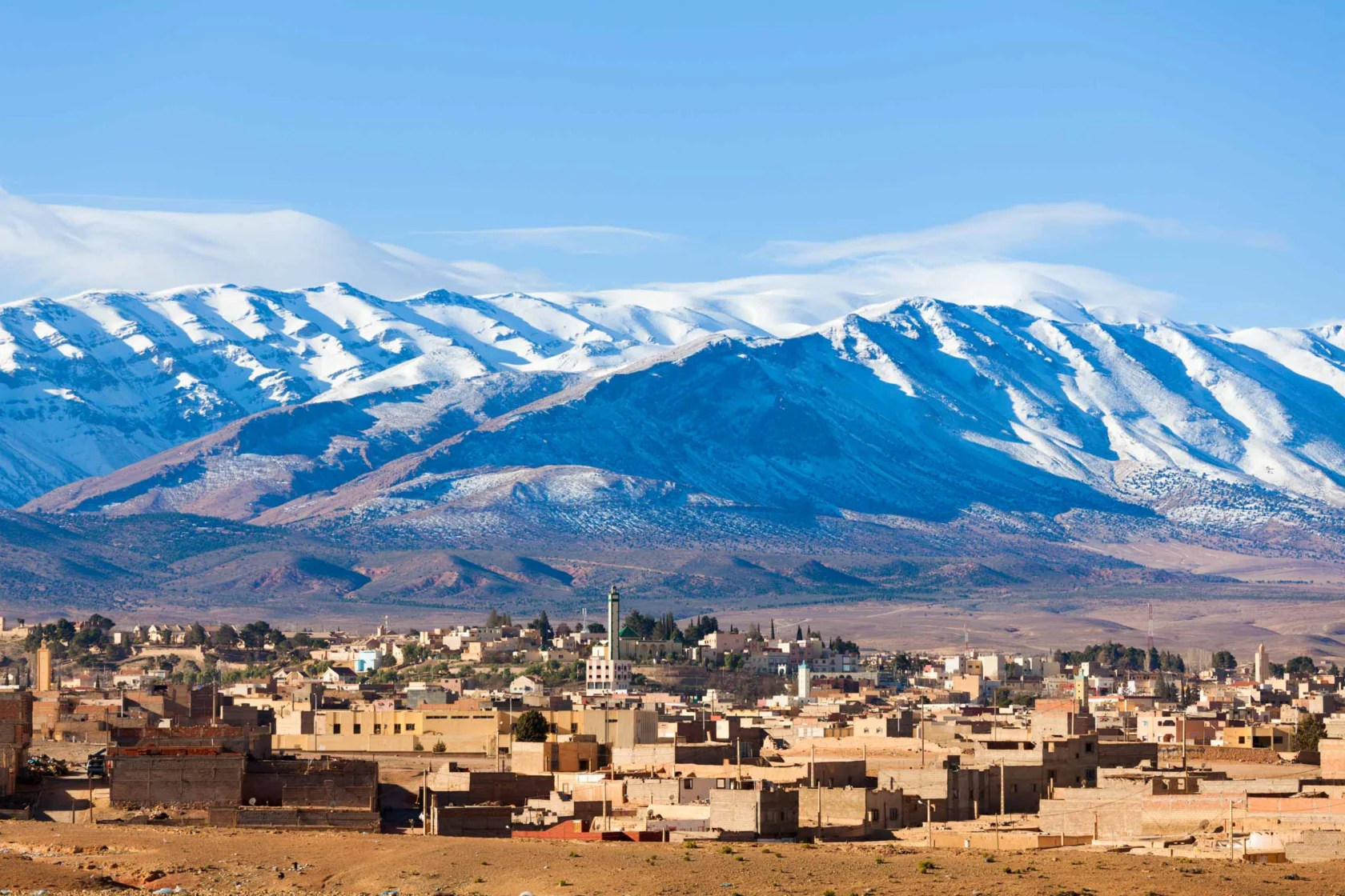 A town with flat-roofed buildings sits in front of snow-capped mountains under a clear blue sky.