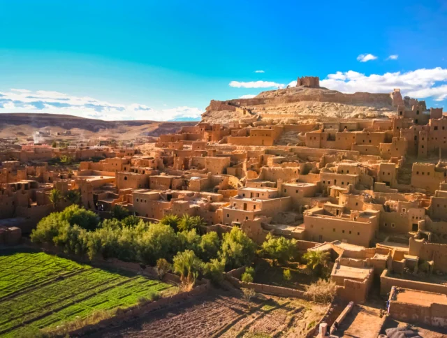 Aerial view of a historic clay village with an ancient fortress on a hilltop, surrounded by desert landscape and green fields under a clear blue sky.