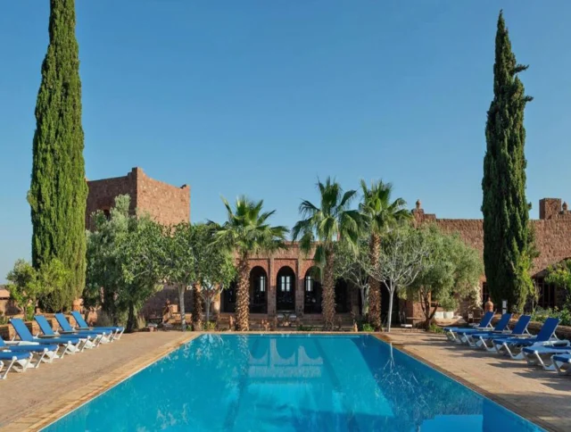 Rectangular swimming pool surrounded by lounge chairs, palm trees, and two tall cypress trees, with a rustic stone building in the background under a clear blue sky.