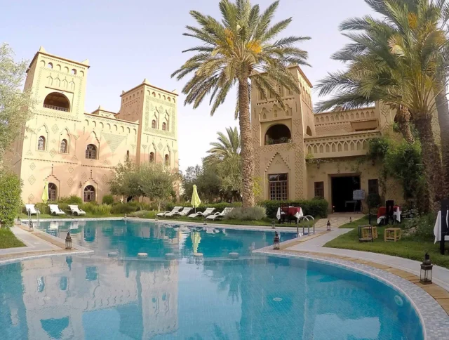 Luxurious poolside view of a Moroccan-style hotel with ornate buildings, surrounding palm trees, and sun loungers.