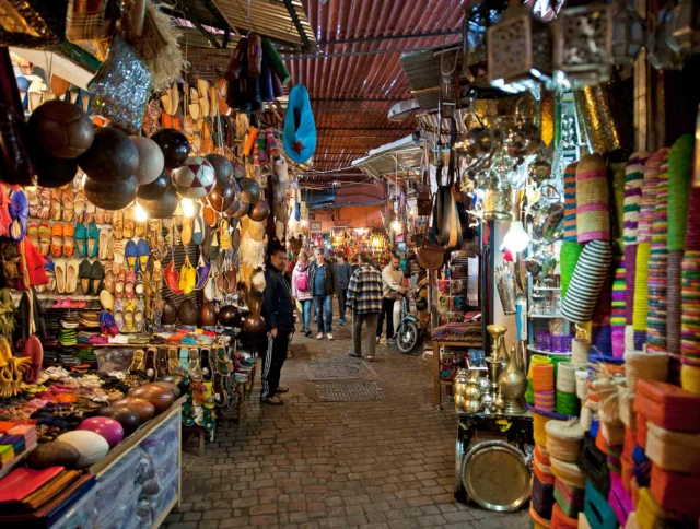 A bustling market scene with colorful textiles, shoes, and crafts displayed. Shoppers browse the vibrant stalls, while some people walk through the narrow aisle.