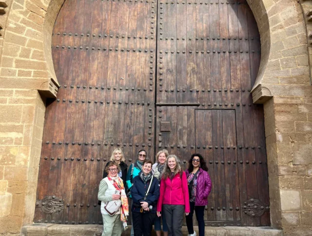 Six people standing in front of a large, ornate wooden door with arches, set in a stone structure.