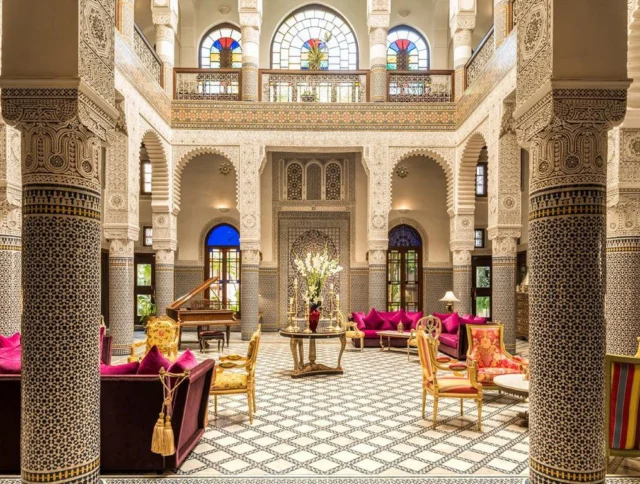 Luxurious hotel lobby with ornate columns, patterned tile floors, arched windows, and vibrant seating areas featuring pink and red cushions. A grand piano is visible in the background.