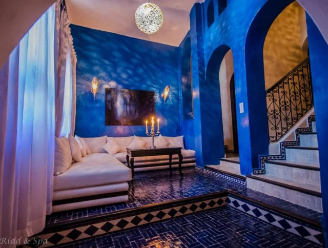 A room with deep blue walls features a white sectional sofa, a table with candles, and a staircase with ornate railings. A spherical light fixture hangs from the ceiling.