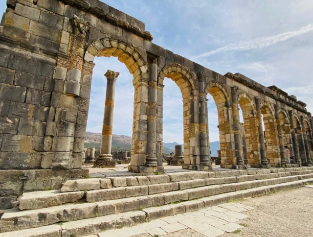 Ancient stone arches and columns, part of a historic ruin with steps in the foreground and a mountain backdrop under a partly cloudy sky.