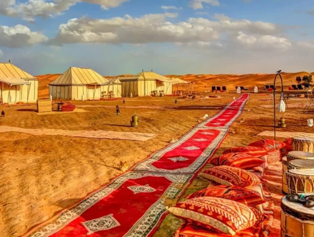 Desert campsite with large tents, a long red carpet, and cushions set on sandy terrain under a partly cloudy sky.