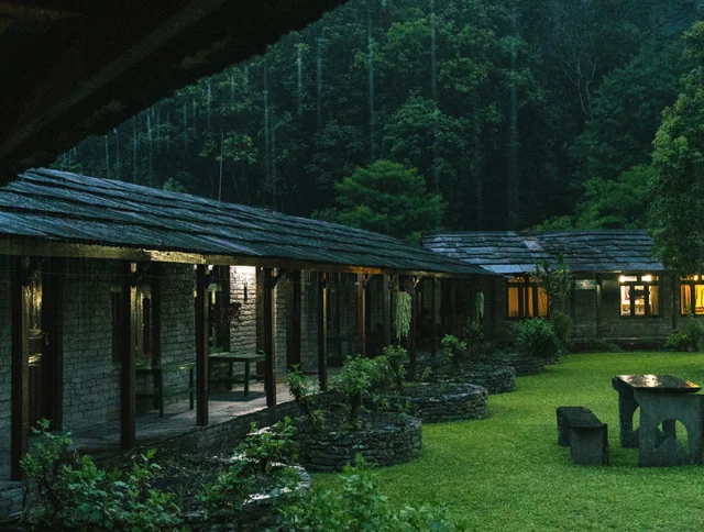 An outdoor courtyard surrounded by stone buildings with wooden roofs and large windows, set in a lush, forested area during the evening.