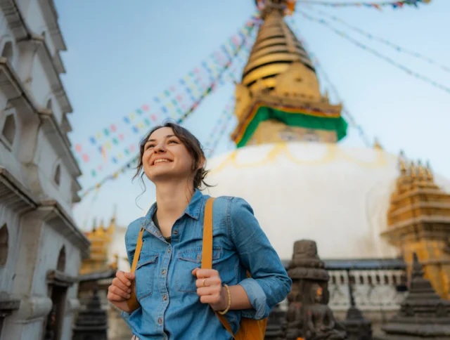 A person in a denim shirt smiles while standing in front of a large stupa adorned with colorful prayer flags.