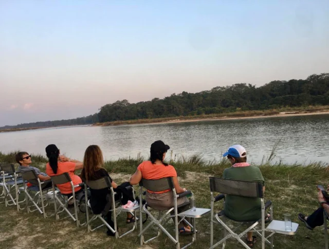 People sit in folding chairs facing a river, with trees in the background, under a clear sky.