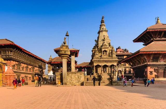 A lively public square with traditional Nepalese architecture, featuring temples and shrines. People are walking and gathering in the area.