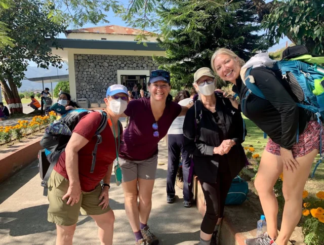 A group of four people wearing casual hiking attire stand smiling outdoors near a building and some trees. Two of them are wearing face masks.